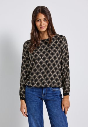 Frau, die eine langärmlige Bluse mit schwarzem und beigem Muster und blauen Jeans trägt, steht vor einem einfarbigen hellgrauen Hintergrund.