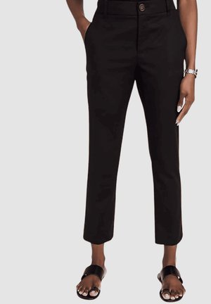 Mujer con pantalones negros cortos con bolsillos, sandalias negras abiertas y un reloj de pulsera en la muñeca izquierda.