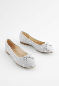 Hopeasa ballet-kenkiä, jotka on valmistettu teksturoidusta kankaasta ja joissa on kimaltavia koristeita sekä koristeellinen iso solki varpaassa. Kenkäparissa on matala korko.