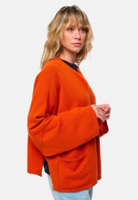 Pull en laine orange surdimensionné avec de longues manches larges, poche frontale et col rond, superposé sur une chemise bleu marine.