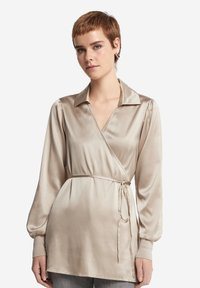 Beige satin omslagsblus med långa ärmar, en spetsig krage och knyt i midjan. Har en slät textur och en subtil lyster.