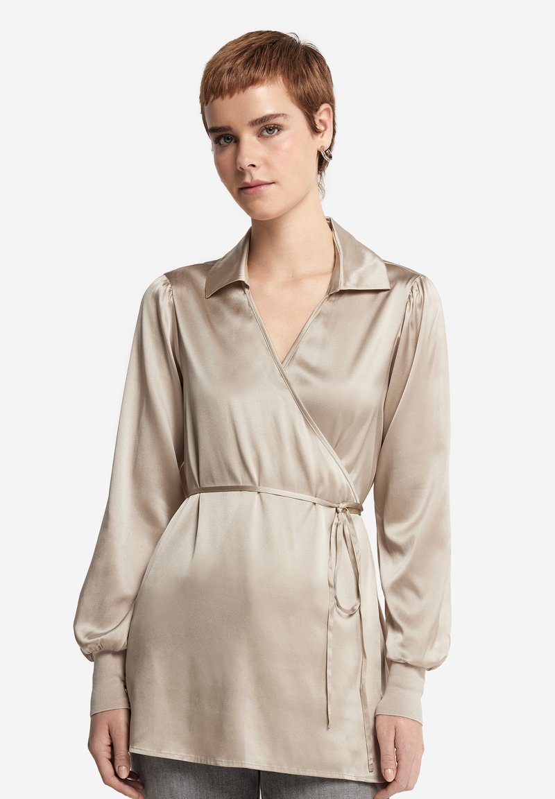 Beige satin omslagsblus med långa ärmar, en spetsig krage och knyt i midjan. Har en slät textur och en subtil lyster.