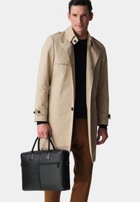 Black leather briefcase with textured finish, dual handles, and zip pocket. Model wears a beige trench coat and brown trousers.