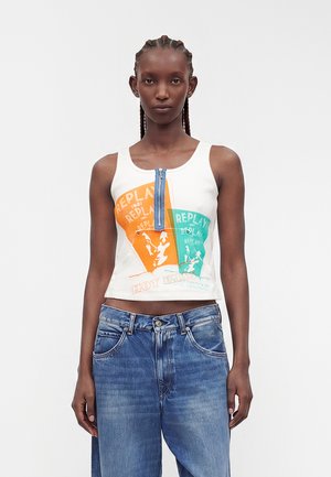 Femme aux cheveux tressés porte un crop top blanc sans manches avec des motifs orange et bleu sarcelle et une fermeture éclair bleue à l’avant, associé à un jean bleu ample.