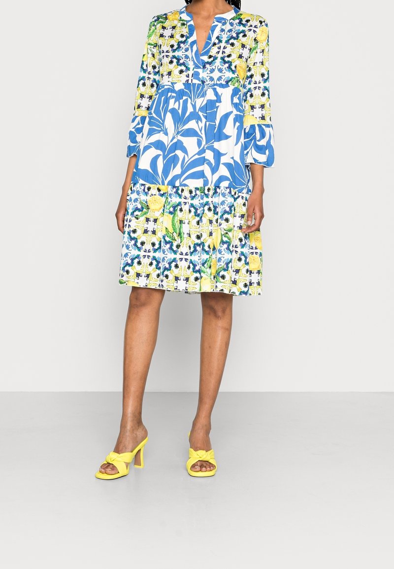 Vestido de estampado floral con una gama de colores azul, amarillo y verde. Presenta un diseño en capas, mangas largas y un escote con cuello. Sandalias de tacón alto rosas.