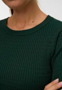 Pull en tricot vert avec un motif texturé en côtes et un col rond classique. Gros plan sur les détails du tissu et de la manche.