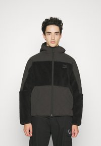 Veste noire matelassée avec un panneau en fausse fourrure noire duveteux, fermeture éclair sur le devant, capuche et manches surdimensionnées. Présente un logo sur la poitrine.