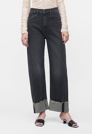 Personne portant un jean noir à jambes larges avec de grands revers retroussés et des sandales noires ouvertes à talons bas, associé à un haut beige clair à manches longues froncé.