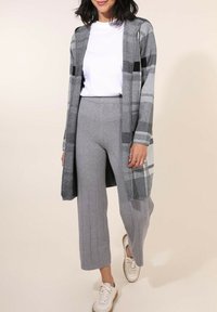 Cardigan à carreaux gris avec des manches longues, associé à un pantalon large en maille côtelée gris et des baskets blanches, sur un fond neutre.