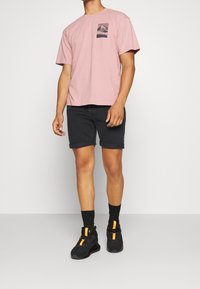 Ljusrosa bomulls-T-shirt med en bröstgrafik, i kombination med svarta denimshorts. Skorna har svarta och orange detaljer.