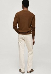 Uomo che indossa un maglione marrone a maniche lunghe, pantaloni avorio e sandali, in piedi rivolto di spalle contro uno sfondo chiaro e semplice.