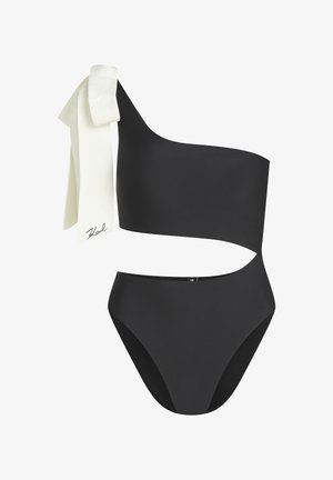 Maillot de bain noir asymétrique avec une découpe à la taille et un nœud blanc noué sur l'épaule, arborant une petite signature.