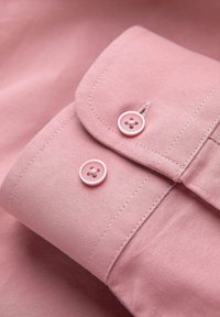 Lichtroze shirtstof met een omgevouwen kraag en twee roze knopen met vier gaten. Vlotte textuur en nette stikdetails.