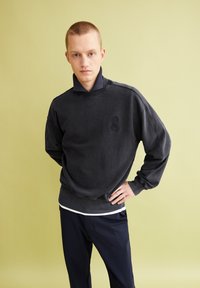 Dark gray sweatshirt with a high collar and a subtle logo. Features dropped shoulders and ribbed cuffs, paired with navy pants.