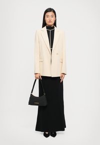Woman standing wearing a beige blazer over a black maxi dress, holding a black Love Moschino handbag with gold lettering.