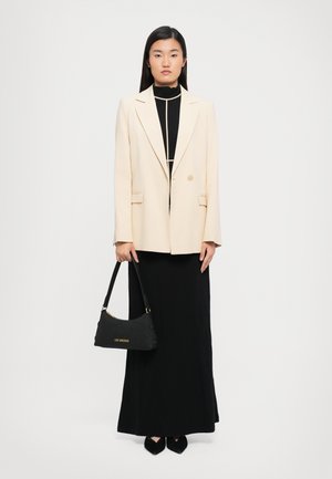 Woman standing wearing a beige blazer over a black maxi dress, holding a black Love Moschino handbag with gold lettering.