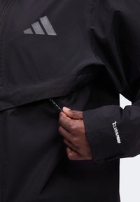 Κλείσιμο μιας φωτογραφίας όπου ένα άτομο ρυθμίζει το φερμουάρ στο μανίκι και το πλαϊνό πάνελ ενός μαύρου μπουφάν Adidas Climaproof.