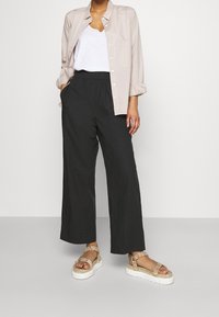 Chemise boutonnée beige, débardeur blanc, pantalon large noir, et sandales à lanières beiges. Design simple avec une coupe décontractée et des textures naturelles.