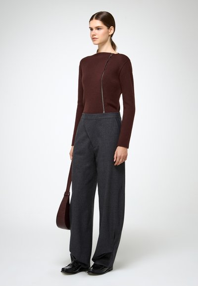 Burgundia pikkade varrukatega kampsun, millel on külgvisiir, koos hallide kõrge vöökohaga laiade pükstega ja mustade kingadega. Mudel hoiab pruuni kotti.