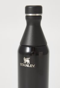 Bouteille d'eau en acier inoxydable noir avec une forme effilée, des accents argentés et le logo "STANLEY" en relief près de la base. Texture lisse, large ouverture.