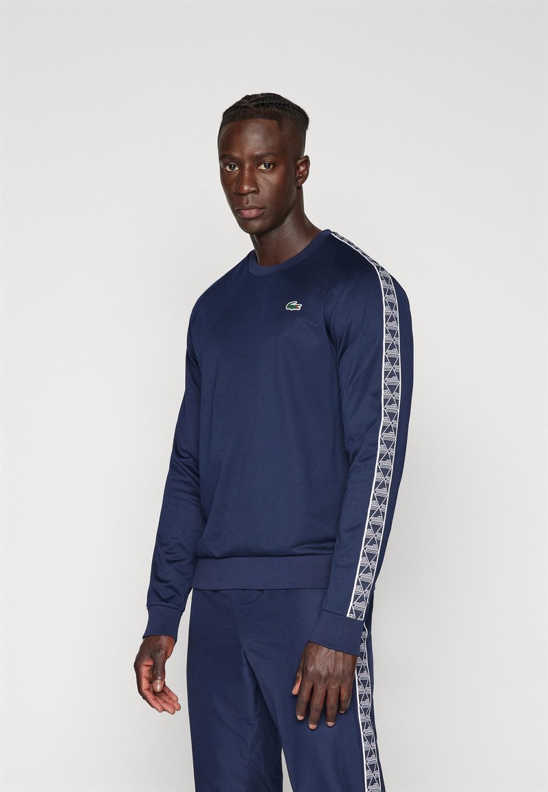 Felpa a maniche lunghe blu navy con strisce bianche a motivo lungo le maniche e un piccolo logo coccodrillo Lacoste sul petto.