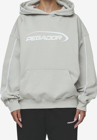 Sudadera gris claro con capucha y logotipo blanco "PEGADOR". Cuenta con puños y dobladillo acanalados, bolsillo tipo canguro y ribetes laterales en contraste.