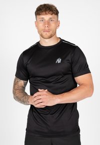 Juodas sportinis marškinėlis, pagamintas iš lygaus, kvėpuojančio audinio. Priekyje yra atspindinčio logotipo ir subtilių akcentų ant pečių. Trumpomis rankovėmis.