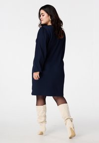 Donkerblauwe gebreide jurk met lange mouwen, losse pasvorm en zijsplitten, gecombineerd met crèmekleurige kniehoge laarzen en doorschijnende zwarte panty's.