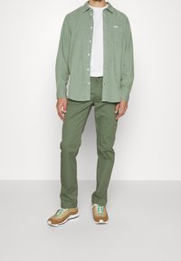 Chemise verte boutonnée au-dessus d'un t-shirt blanc, assortie à un pantalon vert et des baskets marron avec des accents multicolores et des semelles blanches.