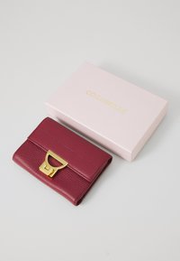 Portafoglio in pelle bordeaux con chiusura in metallo dorato, finitura testurizzata e design a patta, appoggiato accanto a una scatola rosa con branding dorato.