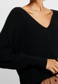 Detailní záběr osoby oblečené v černém žebrovaném svetr s výstřihem do V a dlouhými rukávy, doplněné béžovou sukní s malými černými puntíky.