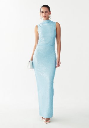 Mujer con vestido largo hasta el suelo sin mangas de color azul claro con lentejuelas, sosteniendo un bolso de mano blanco y llevando tacones blancos de punta abierta y pendientes grandes.