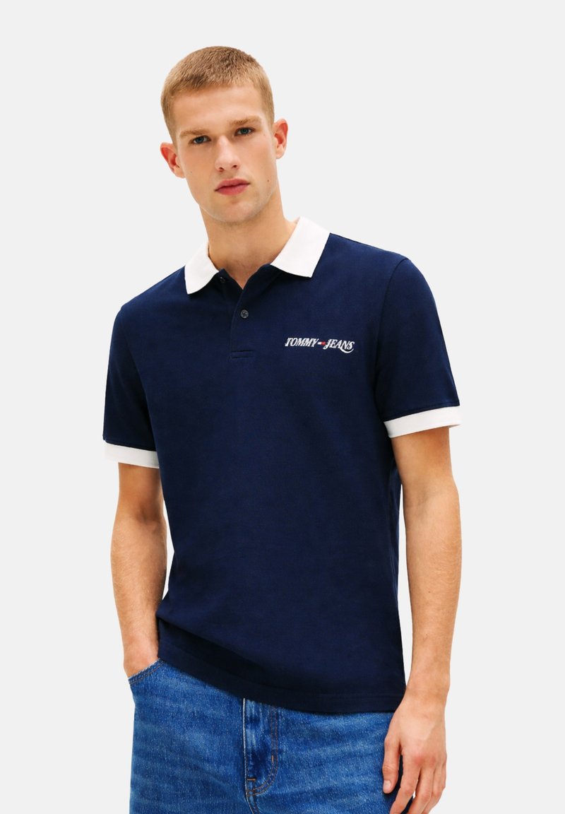 Jeune homme portant un polo bleu marine à col et poignets blancs, logo "Tommy Jeans" sur la poitrine, associé à un jean bleu.