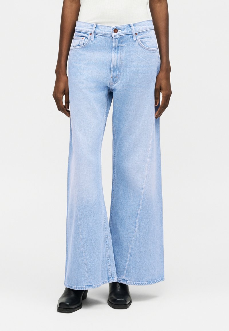 Inimene, kes kannab helehalli laiade säärtega teksapükse, valget tuba ja musta ruudukujulise varbaga kingi, seisab ühevärvilise helehalli tausta ees.