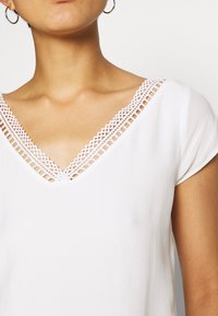 Haut blanc à col en V présentant une bordure en dentelle complexe le long de l'encolure et des manches courtes, fabriqué en tissu lisse et léger.