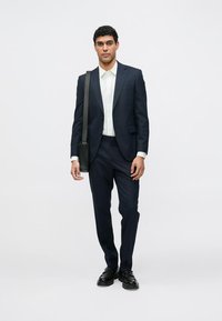 Navy suit with slim fit, textured fabric, white shirt, black shoes, and a black shoulder bag, standing against a plain white background.