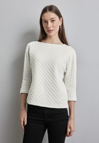 Weißes, gesteppter Langarm-Blusenshirt mit einem Bootsausschnitt und hochgerollten Ärmeln, kombiniert mit schwarzen Hosen. Der Stoff hat ein strukturiertes Rautenmuster.