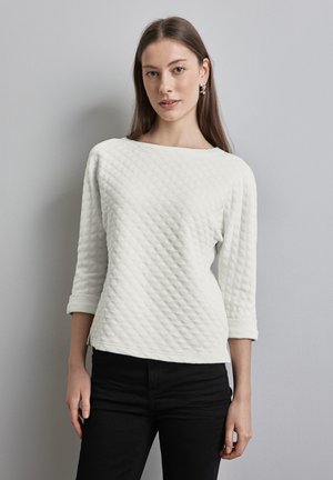 Haut long à manches longues en quilt blanc avec un col bateau et des manches retroussées, associé à un pantalon noir. Le tissu présente un motif en losanges texturé.