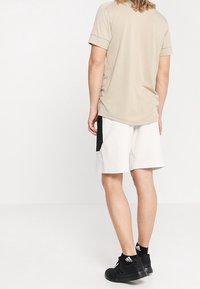 Beige overhemd met korte mouwen en opgerolde mouwen, gecombineerd met witte shorts met een zwart accentzak. Zwarte sportschoenen maken de outfit af.