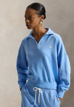 Kvinde med håret bundet tilbage iført lyseblå sweatshirt med lynlås og krave samt matchende bukser med snøre, ser til siden med hænderne i lommerne.