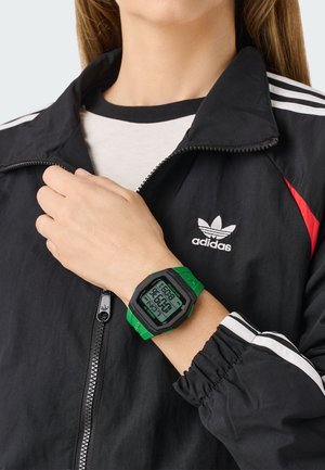 Montre numérique avec un bracelet en silicone vert et un boîtier hexagonal noir, affichant l'heure et diverses fonctions, portée au poignet contre une veste noire.