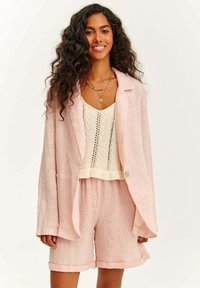 Svetle růžový lněný blejzr s volným střihem, který je zkombinován s krémovým pleteným topem s vzorem chevron, a odpovídajícími růžovými lněnými šortkami.