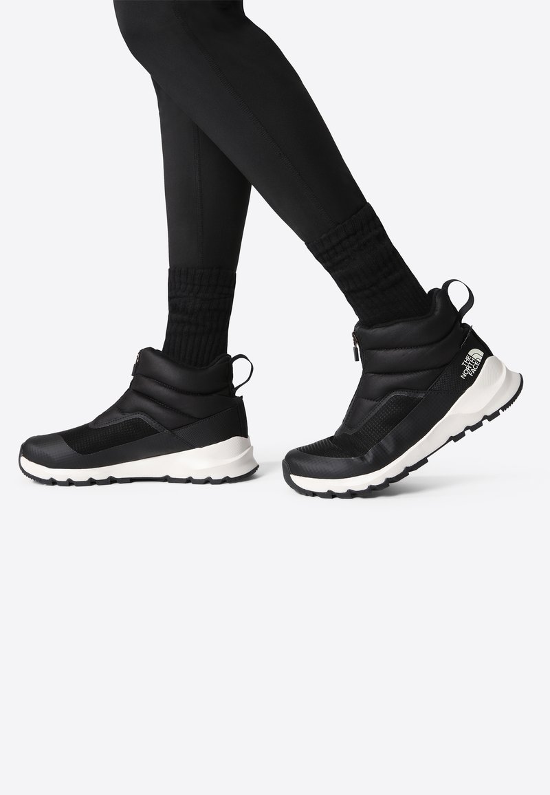 Bottes d'hiver hautes noires avec semelles blanches et fermeture éclair latérale, portées avec des leggings noirs et des chaussettes, montrées en train de marcher à mi-pas.