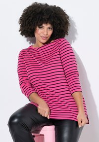 Maglietta a maniche lunghe a righe rosa con righe orizzontali nere, abbinata a pantaloni attillati neri lucidi, modella seduta su uno sgabello rosa.