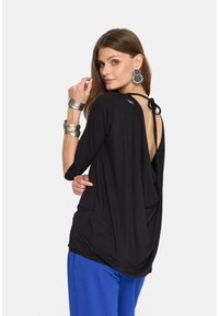 Czarny top z drapowaniem i szerokim otworem na plecach oraz detalem w postaci wiązania. Długie rękawy i miękka tkanina, dopasowany do niebieskich spodni i srebrnych dodatków.