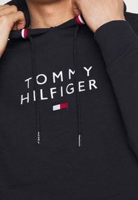 Sudadera negra con capucha que presenta texto bordado en blanco "TOMMY HILFIGER" y un pequeño parche de logotipo en rojo, blanco y azul. Cuerdas de ajuste con puntas de metal.