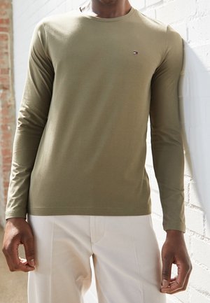 Camisa de manga larga color verde oliva con cuello redondo y pequeño detalle de logo en el pecho, combinada con pantalones de color claro. Textura suave.