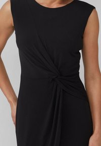 Czarna sukienka bez rękawów, wykonana z miękkiego materiału, z twisted designem w pasie, który tworzy fałdy i elegancko się układa.