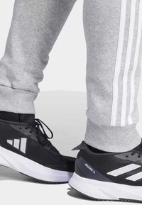 Chaussures de sport noires avec des accents et un logo blancs, associées à un pantalon de jogging gris avec trois rayures blanches sur les côtés.
