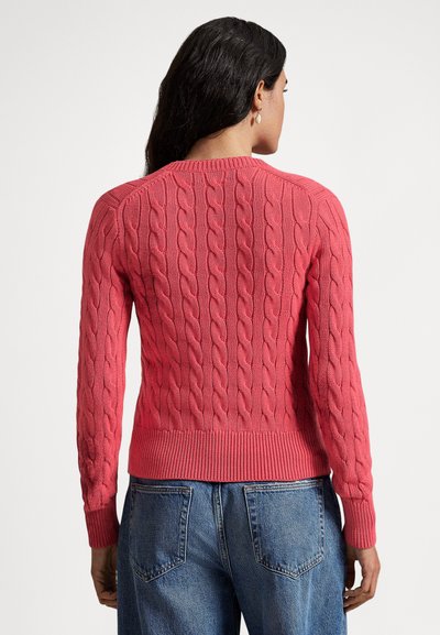 Adīts džemperis spilgtā koraļļu krāsā ar pinuma rakstu, ribām apakšdelmos un apakšmalā, valkāts kopā ar zilām džinsa biksēm. Aizmugurē ir apaļa izgriezuma apkakle.